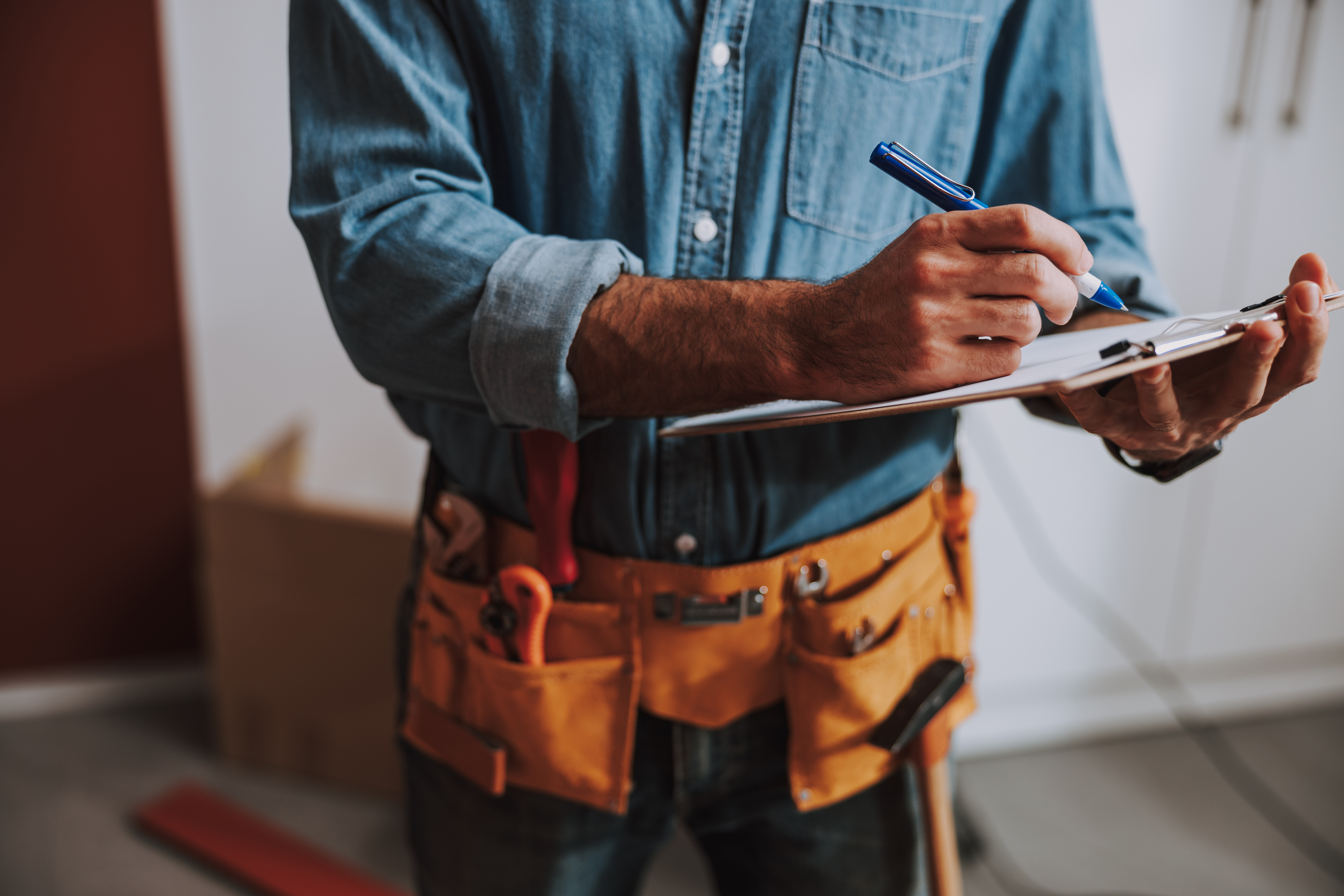 Construction worker in denim shirt and tool belt writing on clipboard with pen